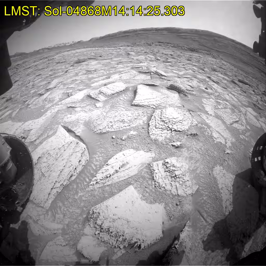 A black-and-white GIF shows the Curiosity Mars rover’s robotic arm drilling into a rock, which lifts up out of the ground. Yellow time stamps in the upper left corner indicate the date and time of each image. The rover’s arm moves around and sand can be seen falling off the rock. The rover’s arm can be seen reorienting the drill more, and finally the rock falls off and fractures. The view, across an expanse of rocks, stretches into the distance with mountains visible in the background.