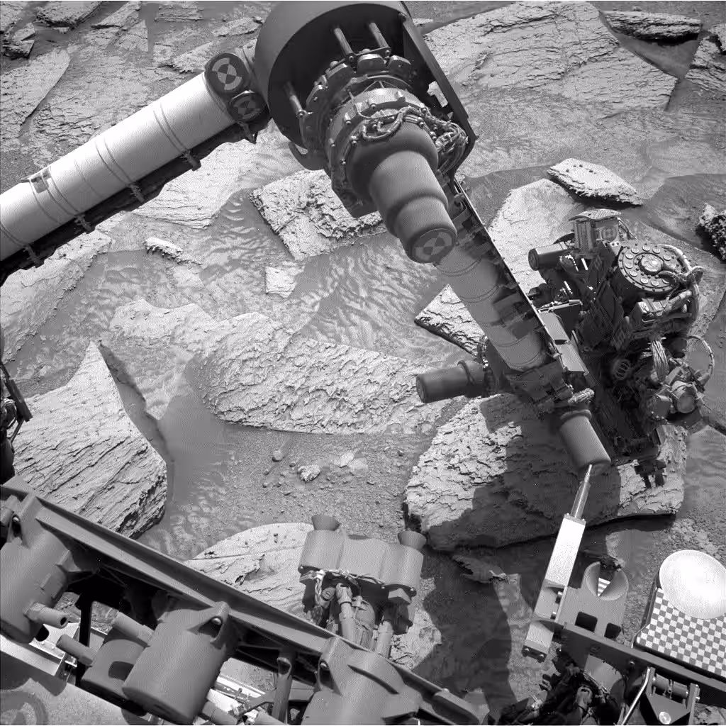 A black-and-white GIF shows the view from the mast, or head, of the Curiosity Mars rover as its robotic arm drills into a rock, then lifts up out of the ground. The rover’s arm moves around and sand can be seen falling off the rock. The rover’s arm can be seen reorienting the drill more, and finally the rock falls off and fractures. The view, across an expanse of rocks, stretches into the distance with mountains visible in the background.