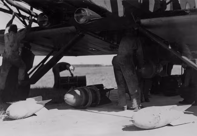 1935 Bombs Black and white photo of three bombs on the ground next to a plane and the back of two people in flight suits