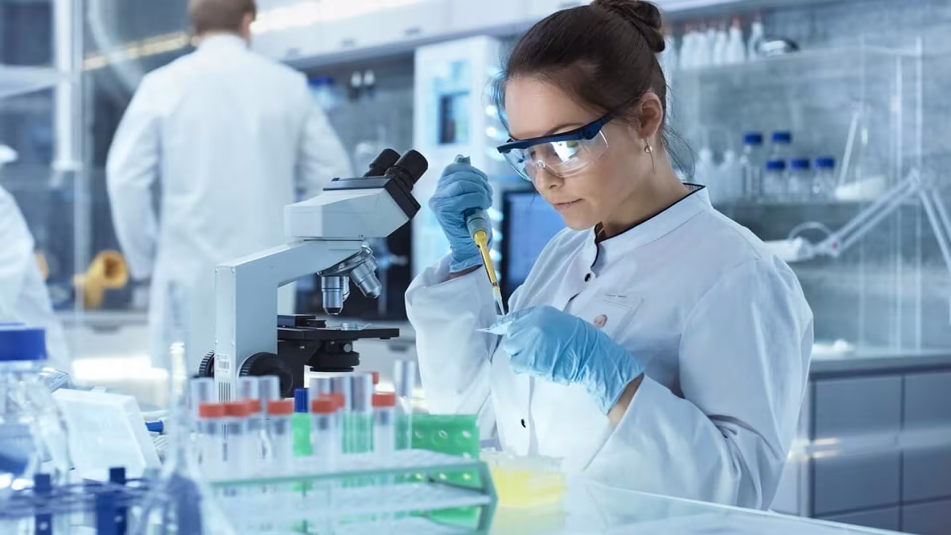 Female Research Scientist Uses Micropipette Filling Test Tubes in a Big Modern Laboratory. In the Background Scientists are Working.