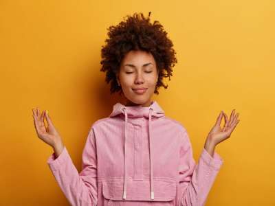 Relieved ethnic woman stands in lotus pose, tries to meditate during quarantine or lockdown, reaches nirvana, does yoga, keeps eyes closed, dressed in sweatshirt.