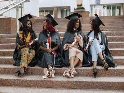 Group of young afro american female students dressed in black graduation gown. campus as a background.