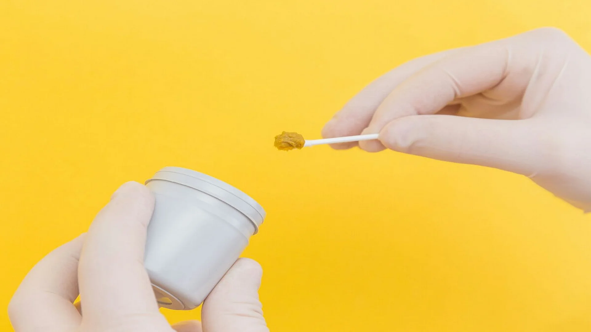 A doctor's hand in a white medical glove holds a stool sample on a stick and a container for stool analysis on a yellow background. The medicine. Analysis. Cala. Infection. The close plan.