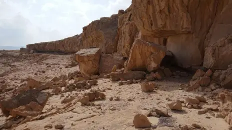 A wide-angle view of a rugged, tan-colored rock cliff and boulders in the Sinai desert under a cloudy sky. The formation includes a low overhang that served as an ancient rock shelter and archaeological site.