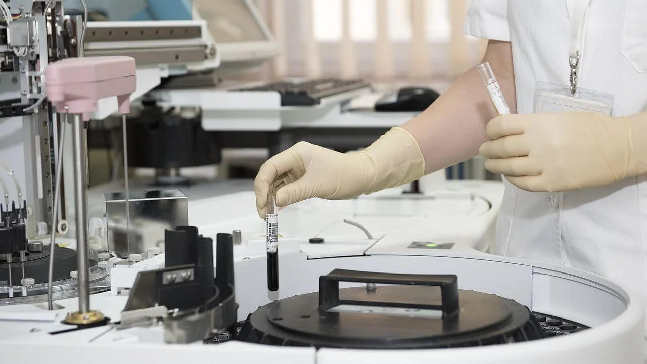 Clinical laboratory professional wearing gloves placing a blood sample tube into an automated chemistry analyzer, illustrating the analytical phase of testing which is subject to clinical audit.