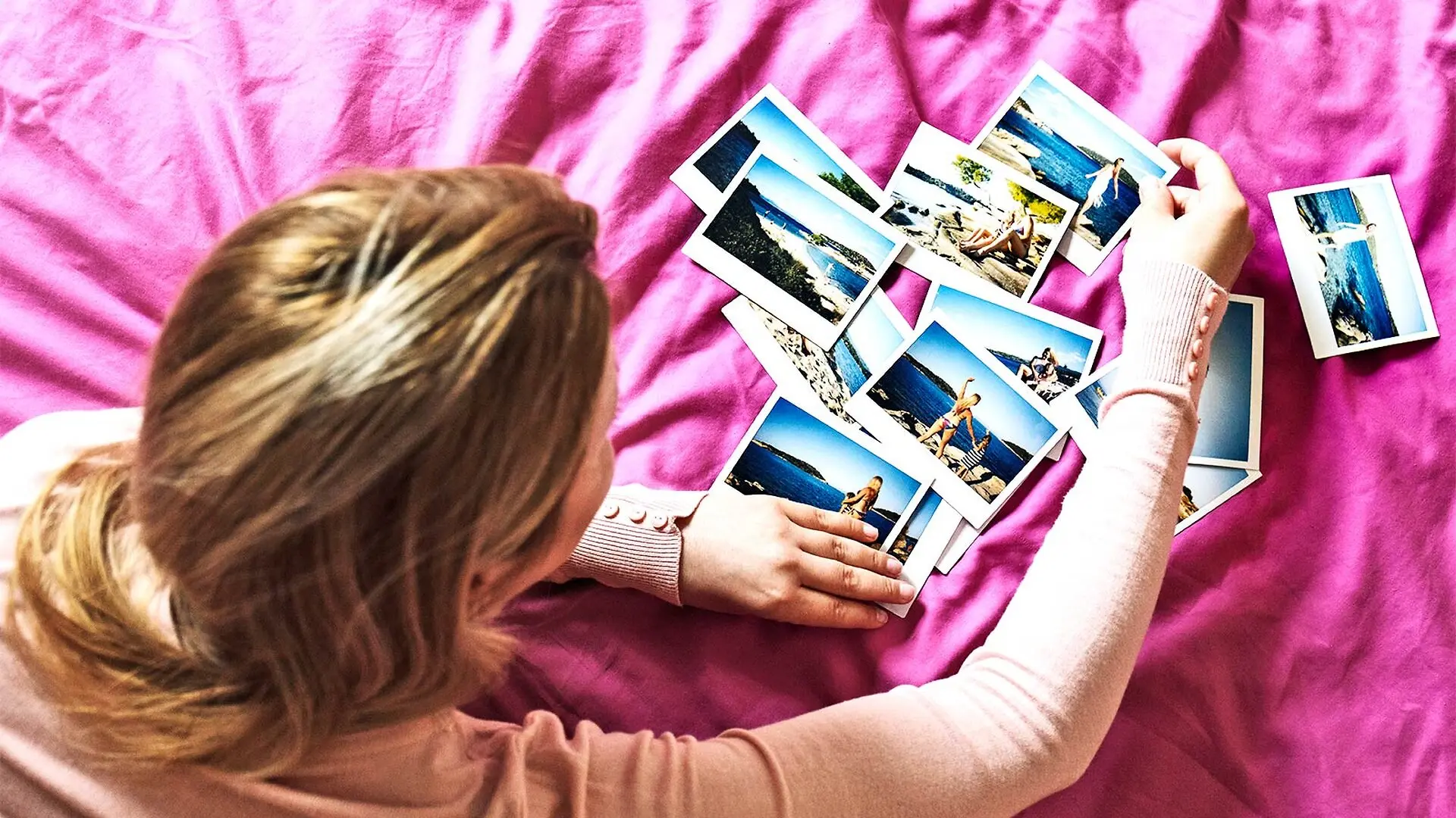 A woman on a pink sheet on a bed looks over photographs.