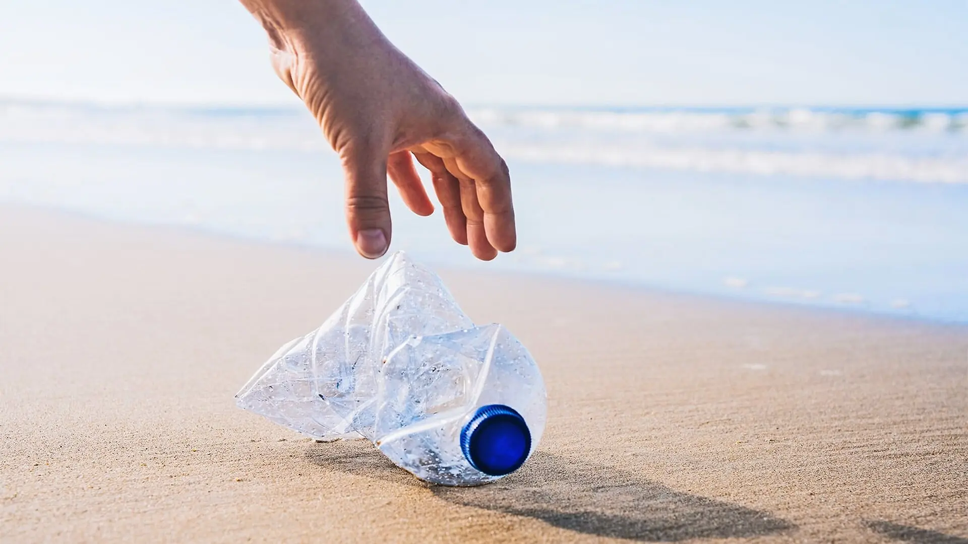 A man reaches for a plastic bottle sitting on a beach.
