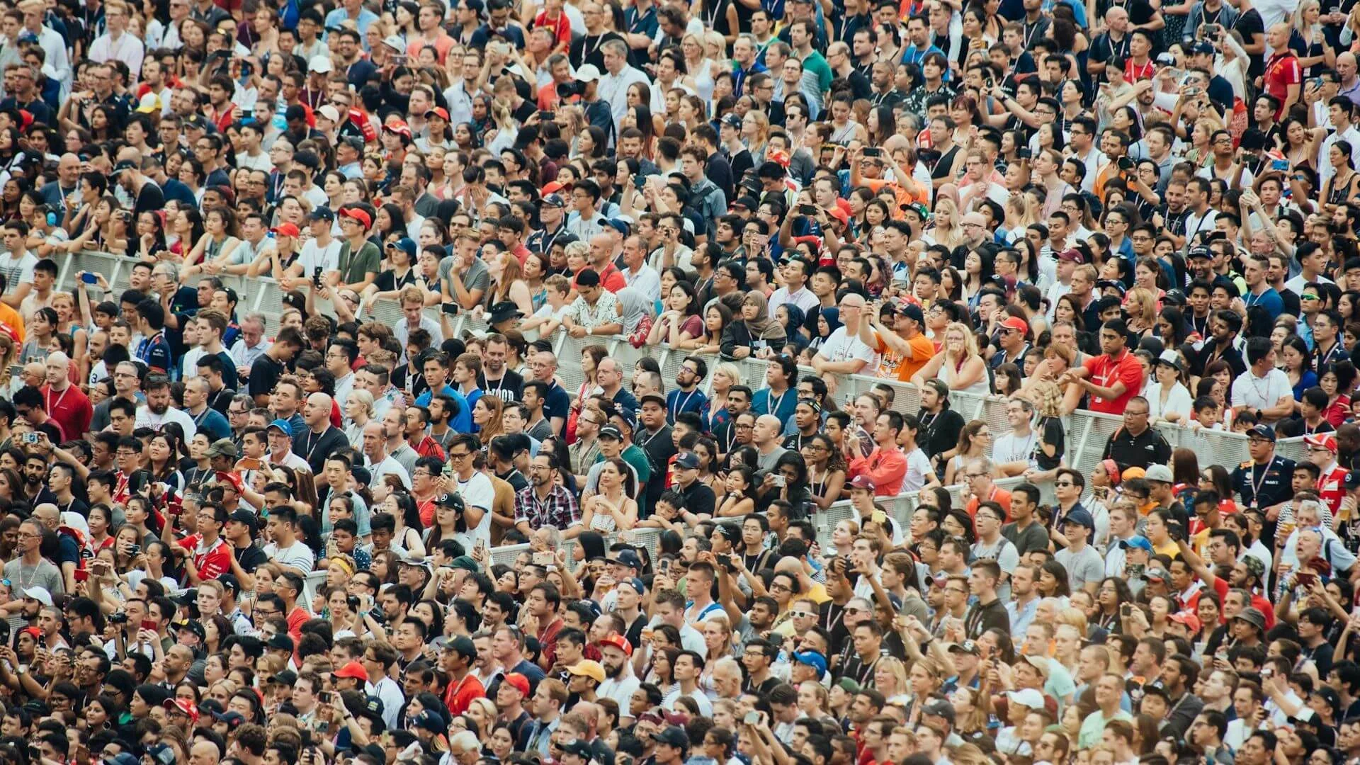 A dense, multi-ethnic crowd of people standing behind a silver metal barrier at an outdoor event, with many individuals holding up smartphones to capture photos or video.