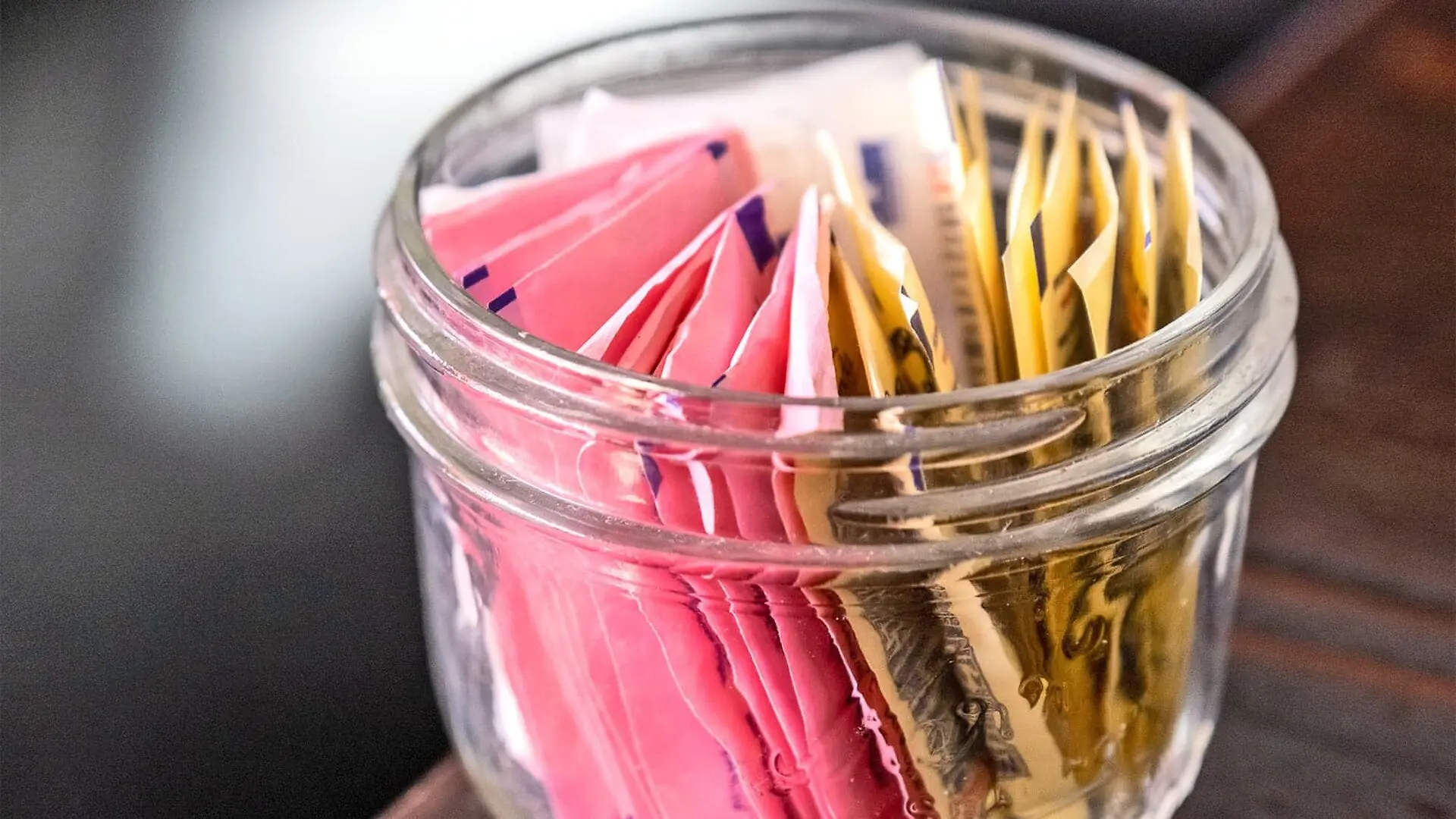 A small jar on a wooden table contains many different packets of artificial sweetener.
