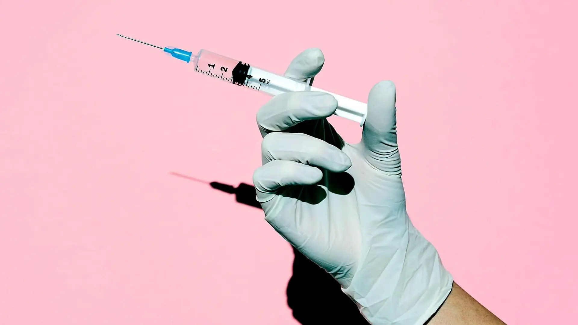 A person wearing a medical glove holds a syringe against a pink background.