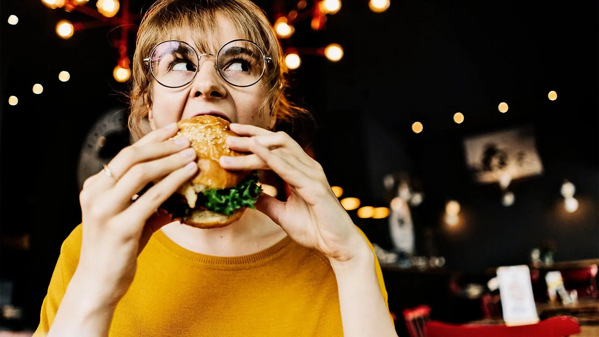A woman wearing glasses takes a big bite of a burger.