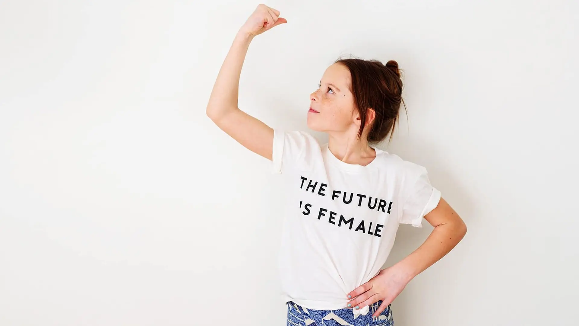 A young girl flexes her bicep while wearing a t-shirt that says 