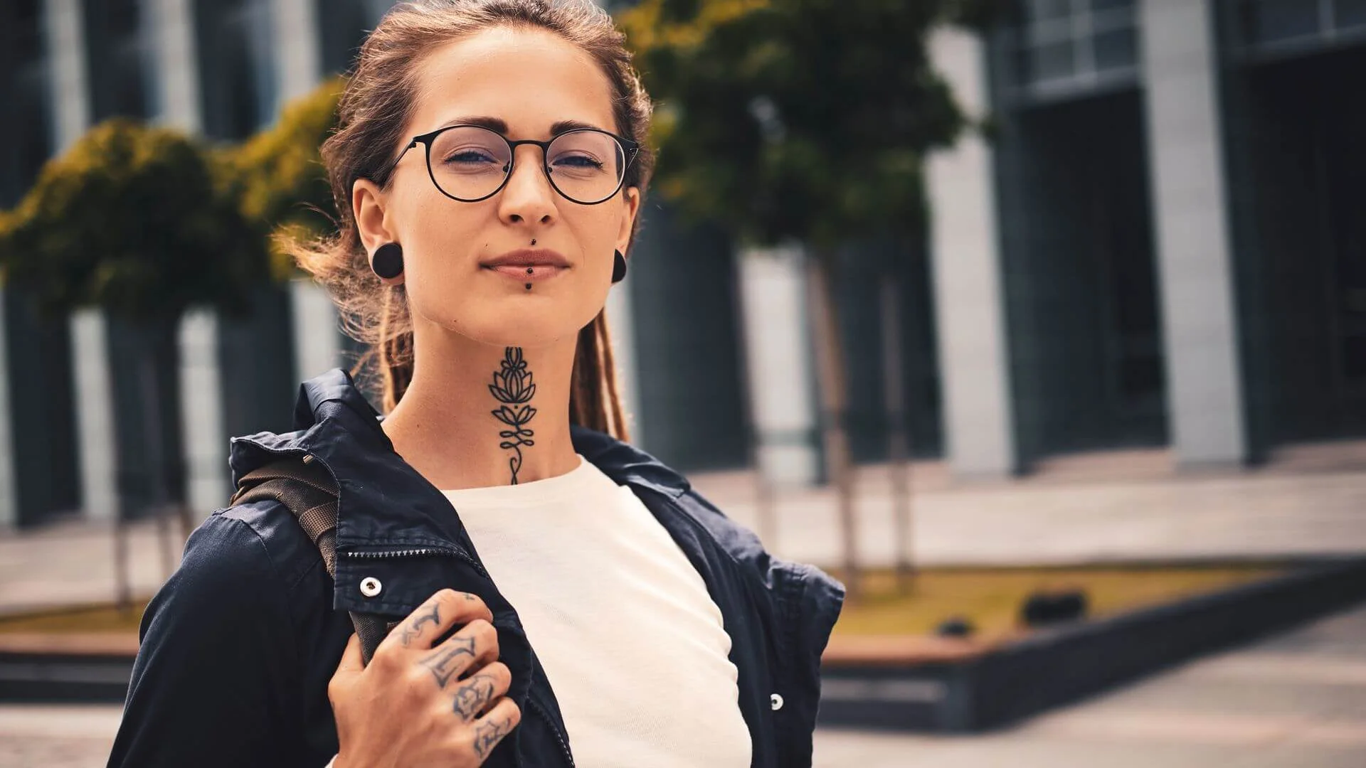A woman with a distinctive floral neck tattoo and several hand tattoos wearing glasses and a dark jacket in an urban setting.