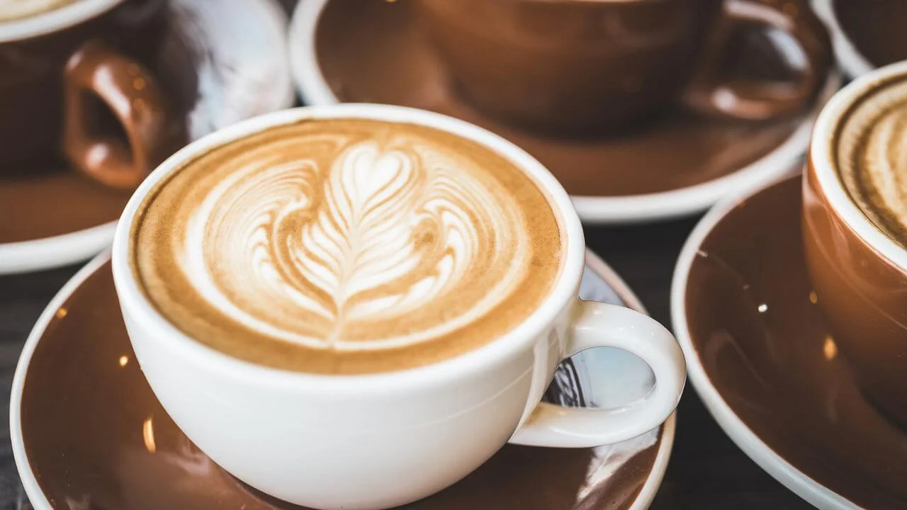 A white ceramic cup filled with coffee featuring detailed latte art in a rosetta pattern, surrounded by several dark brown ceramic cups and saucers.