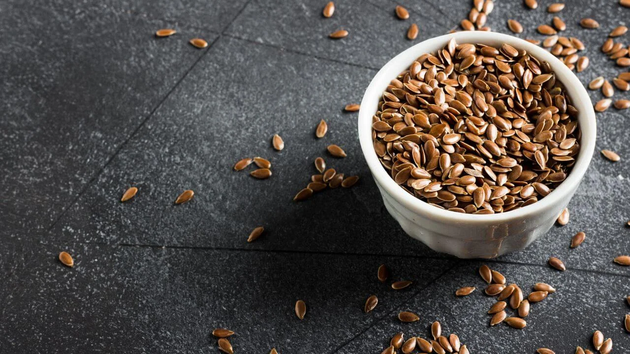 Close-up of whole brown flaxseeds in a small bowl, with some seeds scattered on a dark background.