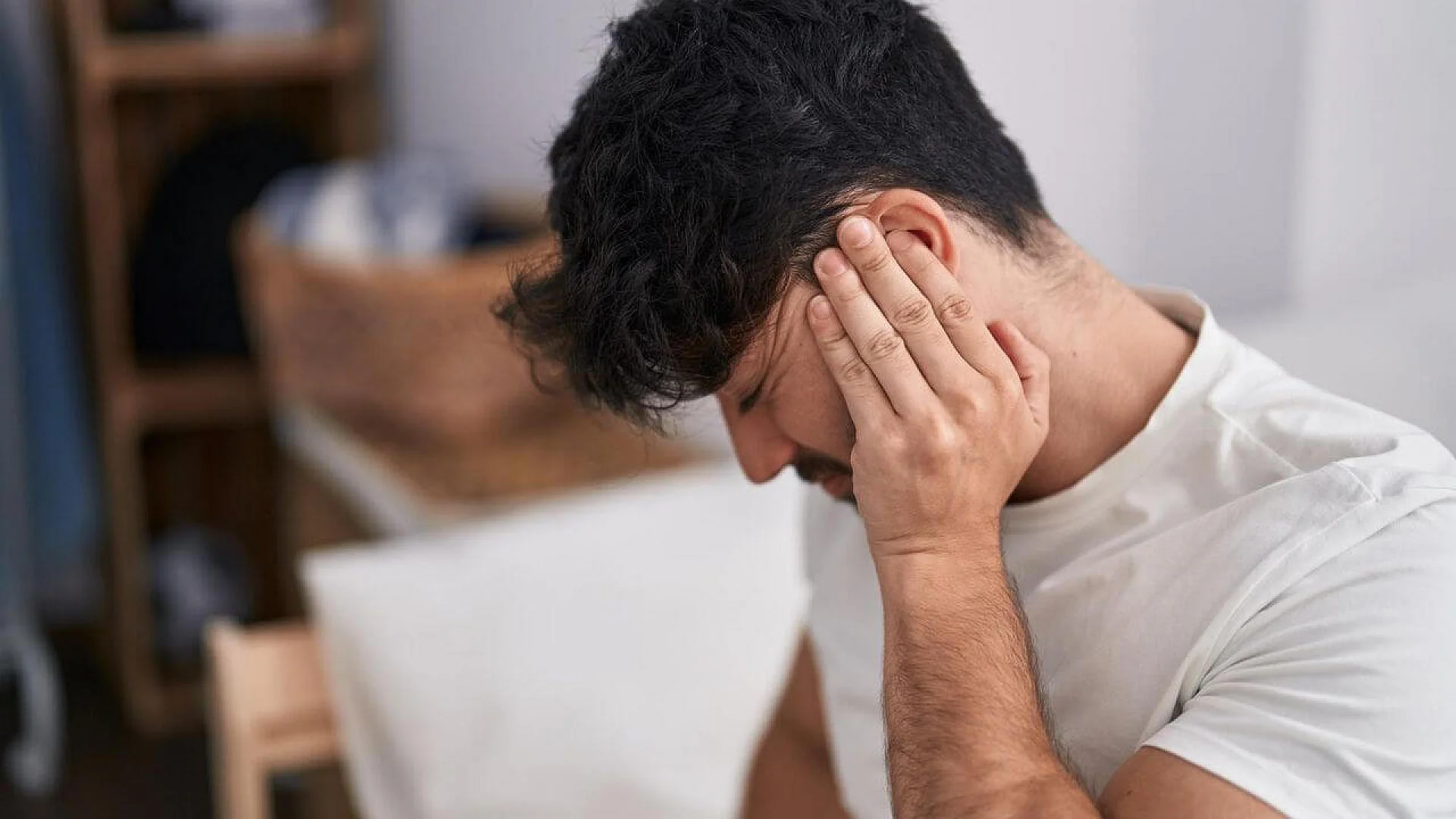 A man in a white shirt sits with his head bowed, pressing his left hand against his ear and temple with a pained expression.