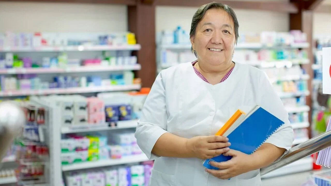 A smiling, middle-aged woman, who appears to be a pharmacist, wearing a white coat and holding notebooks while standing in an aisle of a pharmacy with shelves stocked with various products in the background.