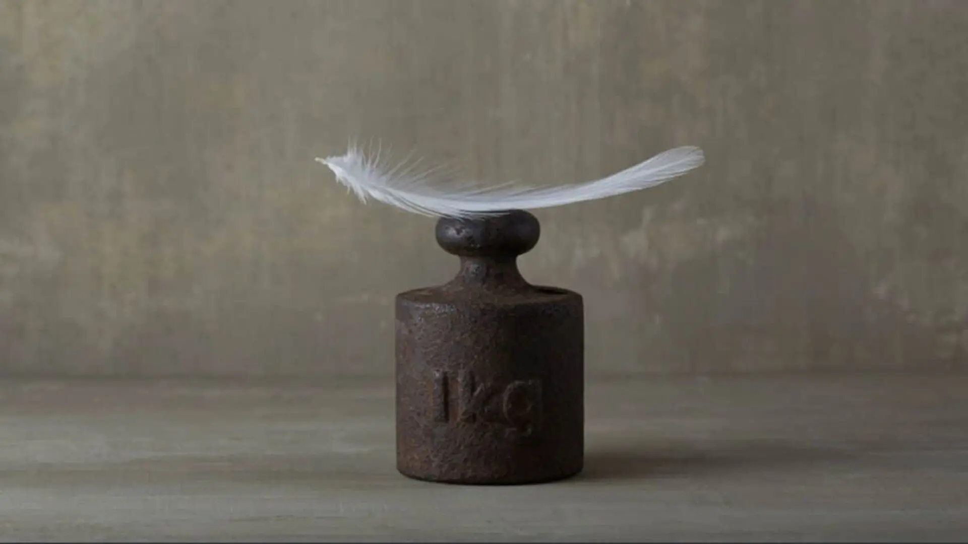 A Vintage One Kilogram Metal Weight With A Long Slender Feather Resting On Top Set Against A Concrete Background Scaled