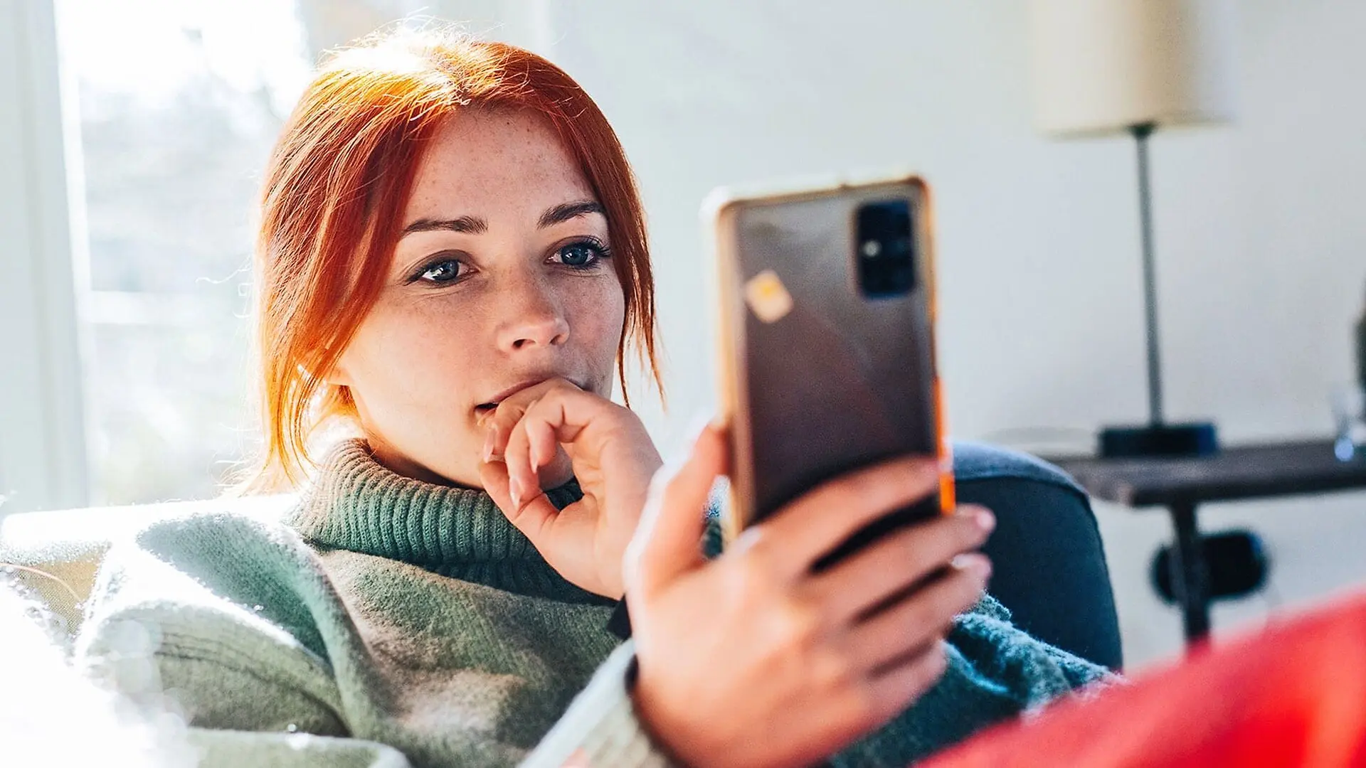 A young woman with red hair reads off of her phone while sitting on a couch.