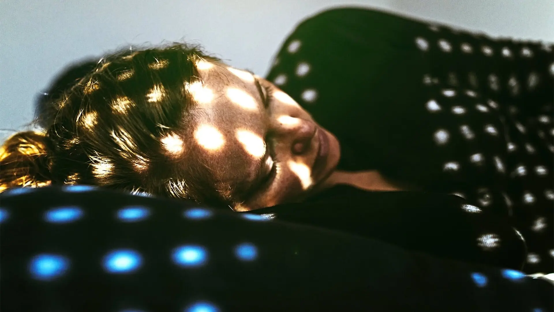 A woman lays in bed while light from outside shines in spots over her face and body.