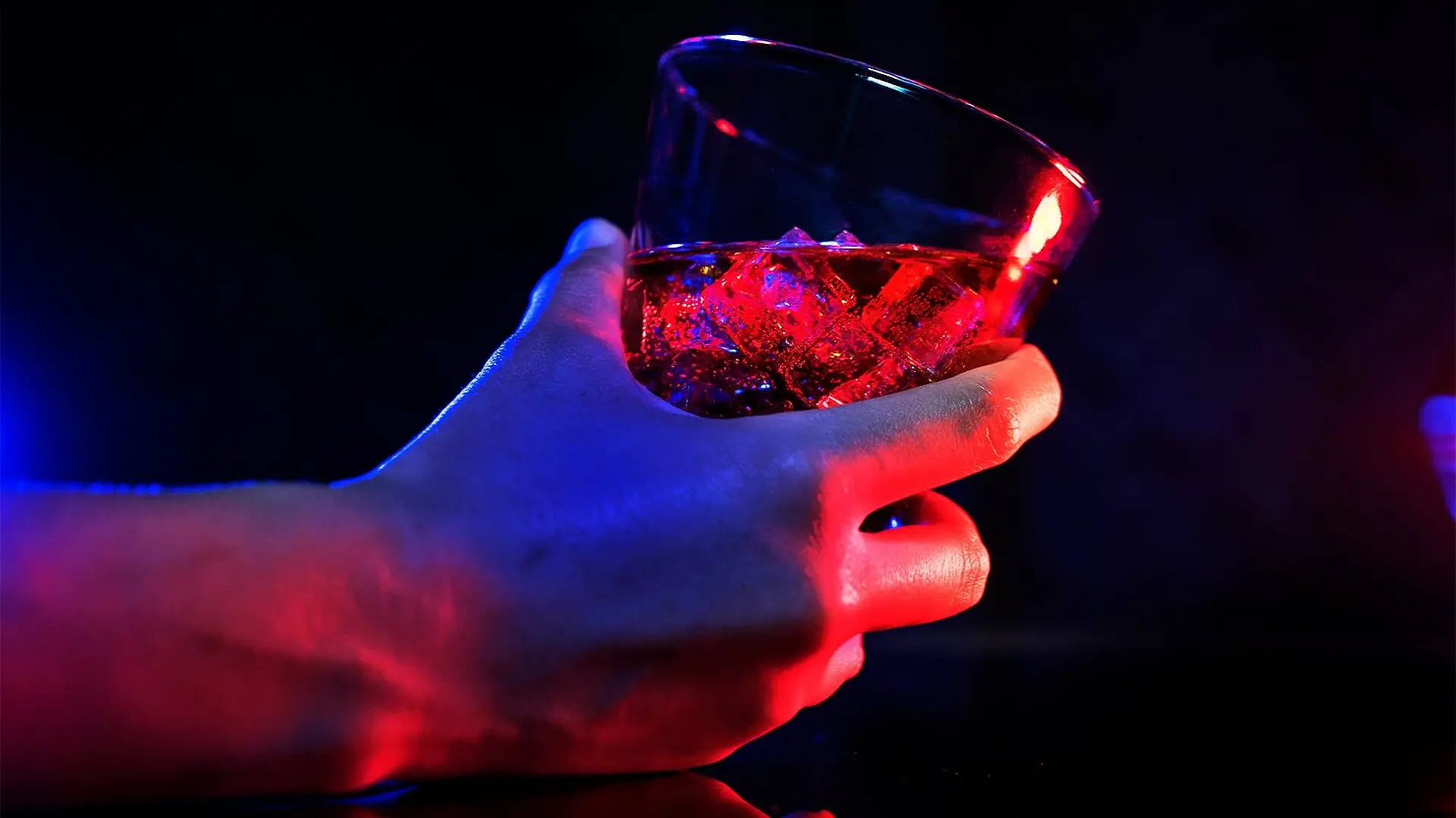 A person holds a glass of alcohol as red and blue light illuminate the scene.