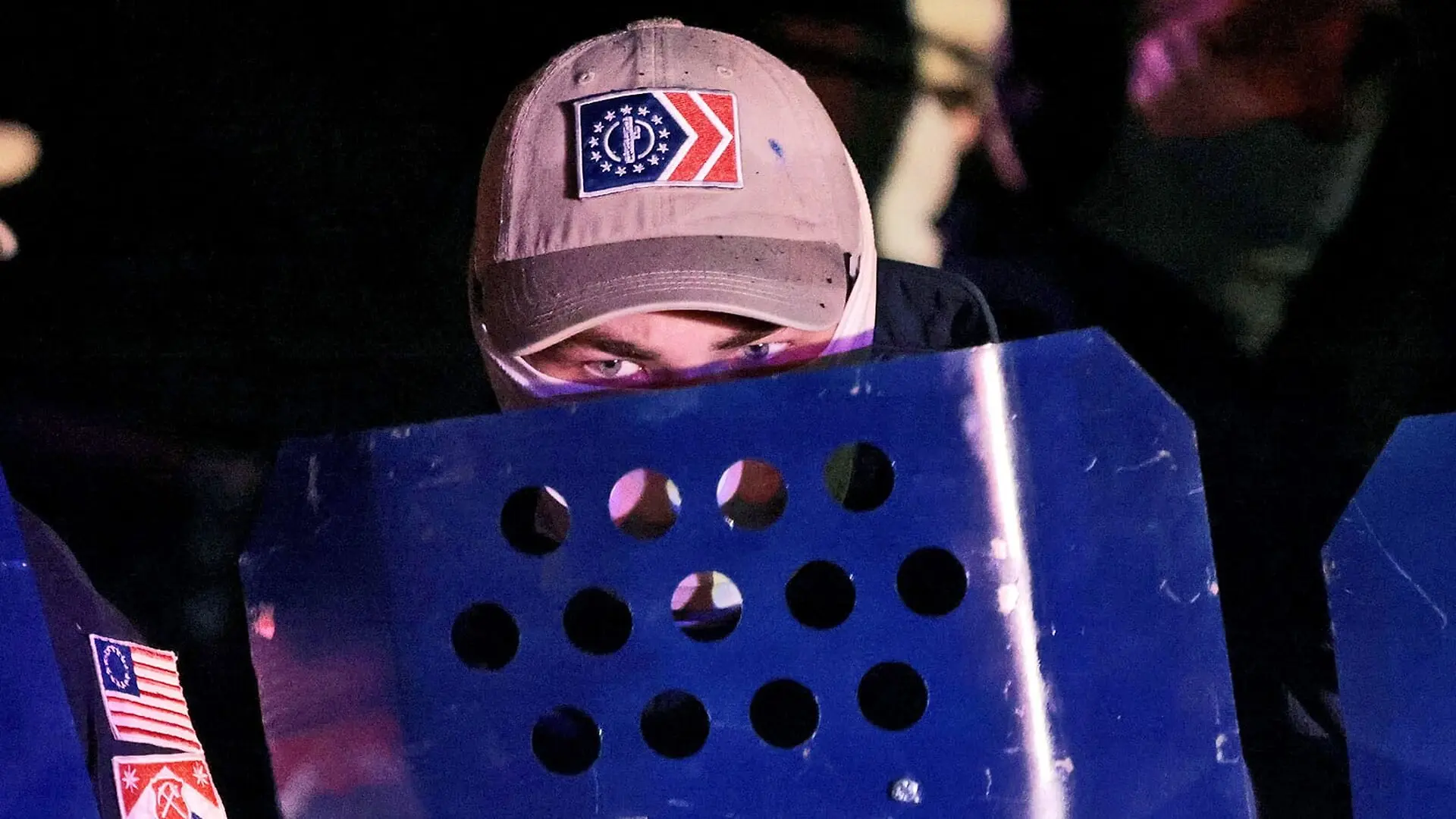 A man hides his face behind a blue shield while wearing a baseball cap.
