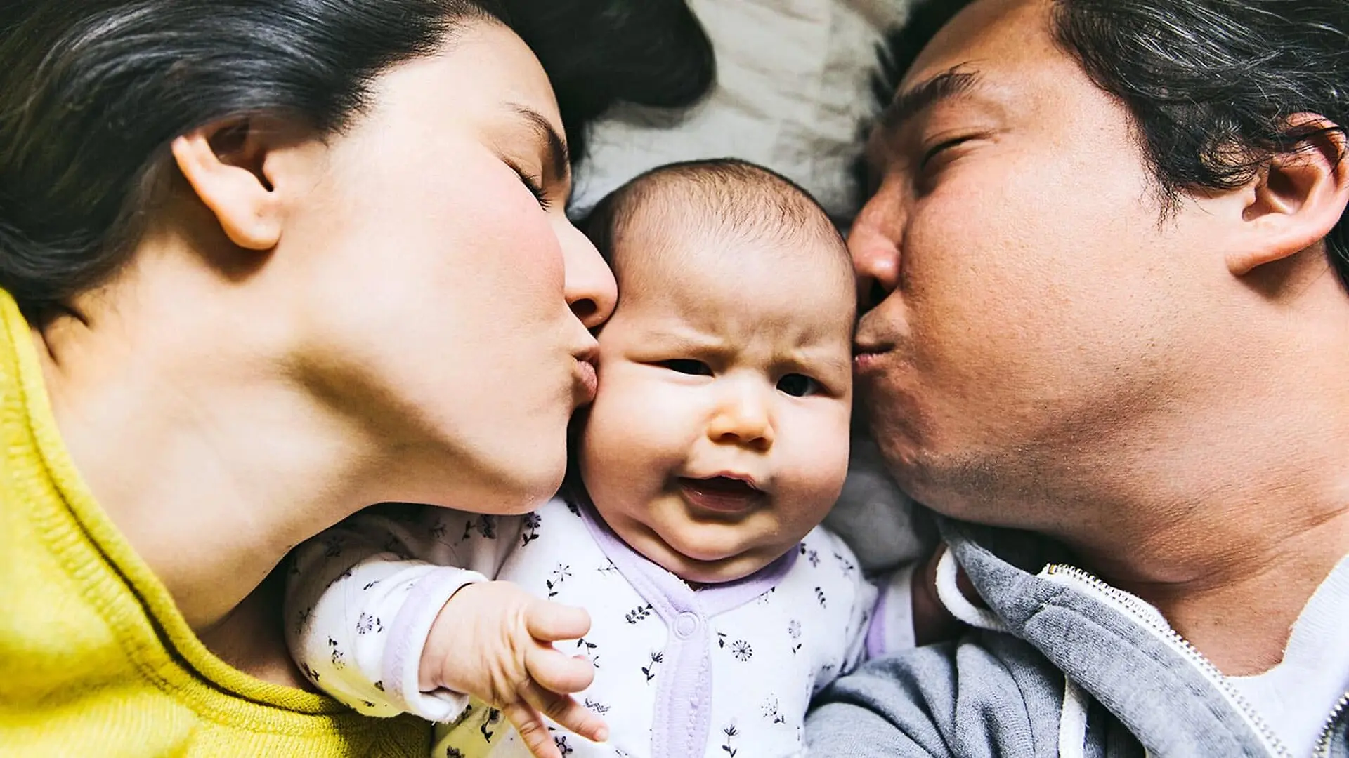 Two parents kiss their baby on either cheek as the baby grimaces.