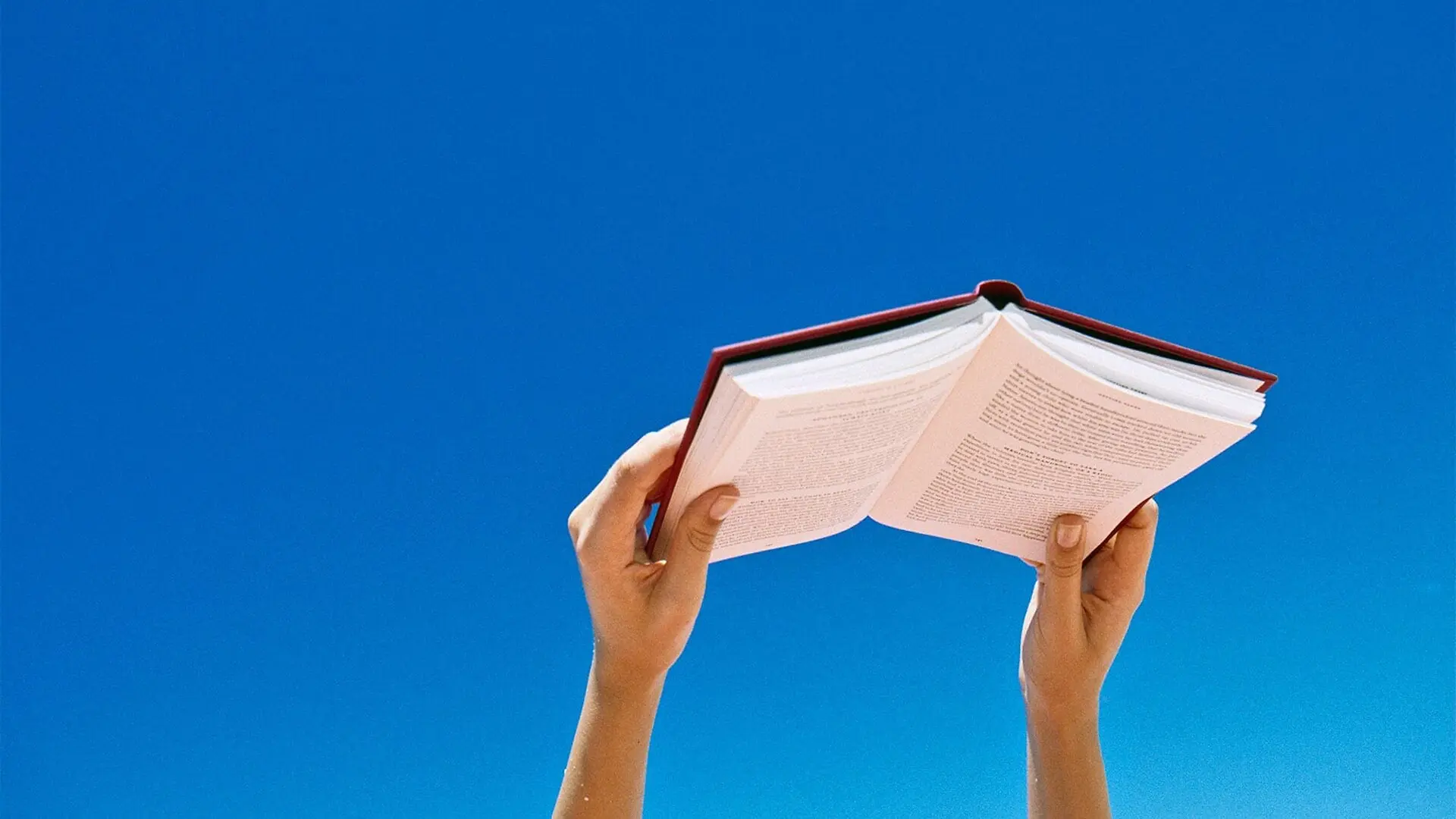 A person holds up a novel against a deep blue sky.