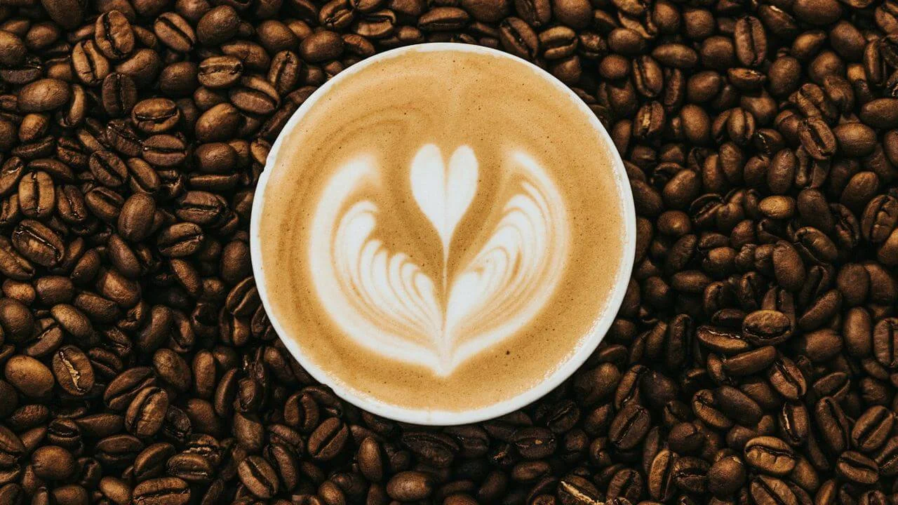 A top-down view of a white ceramic cup containing a latte with heart-shaped foam art, centered and resting on a dense bed of roasted brown coffee beans.