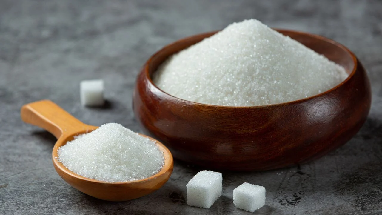 A wooden bowl filled with white granulated sugar alongside a wooden scoop and several sugar cubes on a dark, textured surface.