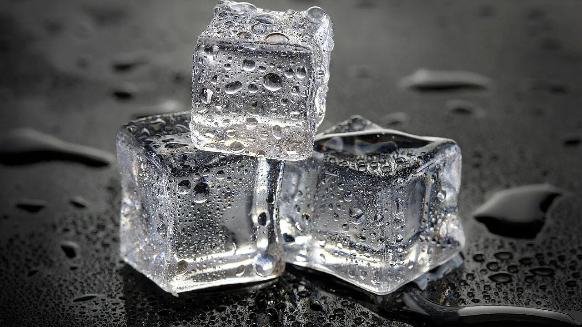 Three clear ice cubes stacked on a dark, reflective surface with scattered water droplets, illustrating the transition between liquid and solid states.