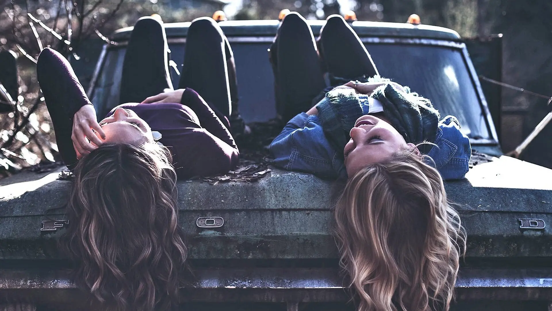 Two teen girls talk while laying on the hood of a car.
