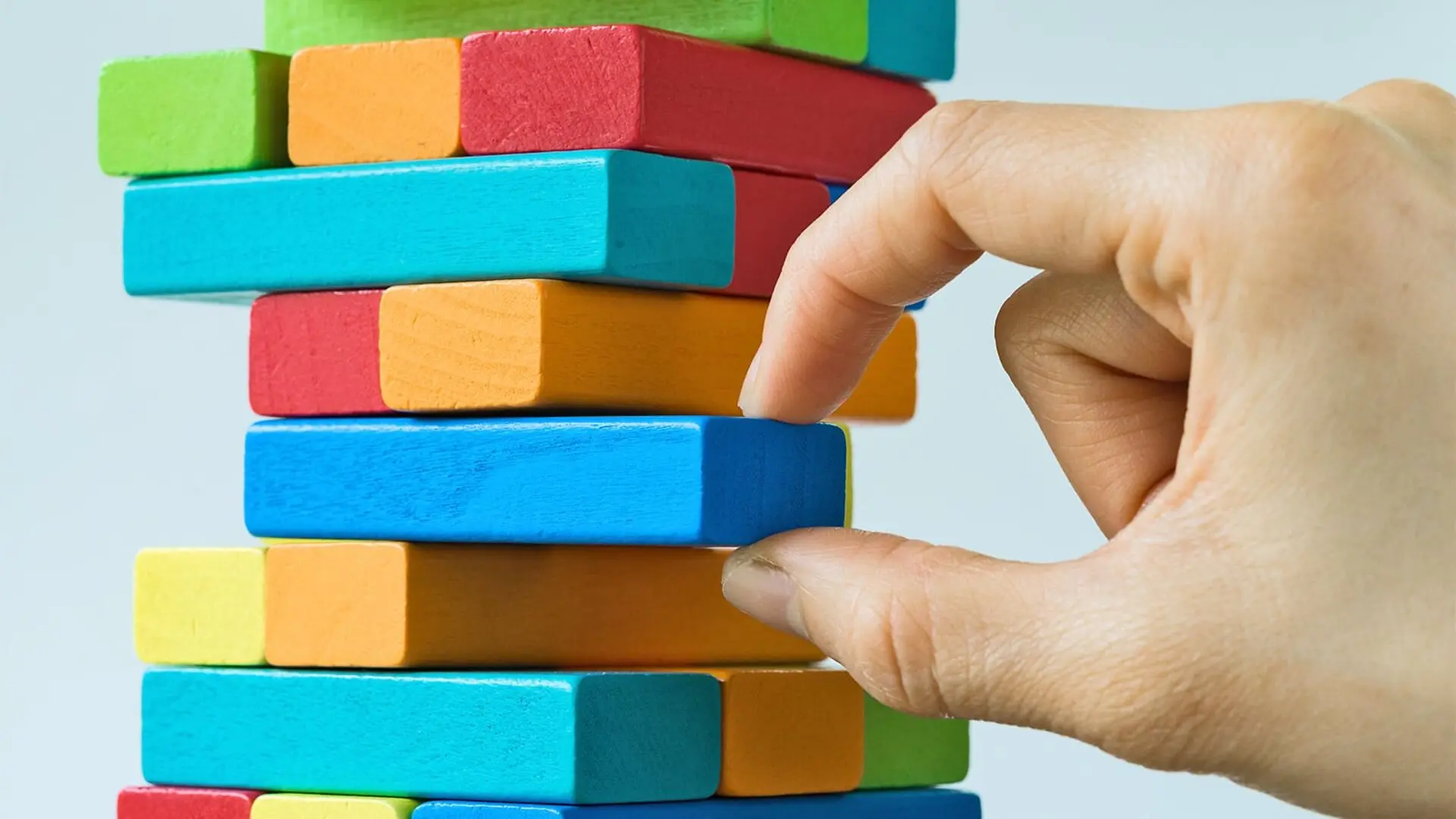 A hand removes one block from a tower of colorful wooden blocks.