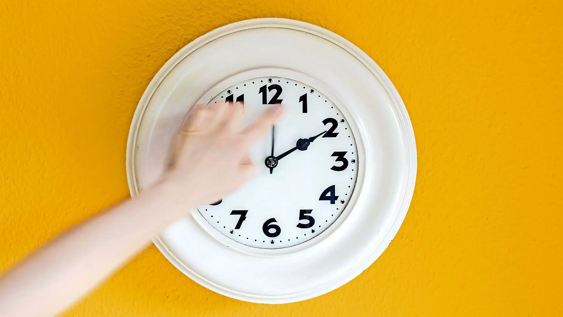 A hand reaches out from off-camera to turn back a white clock on a yellow wall