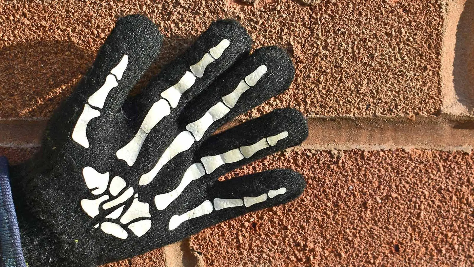A hand with a black glove with skeleton fingers on it touches a brick wall.