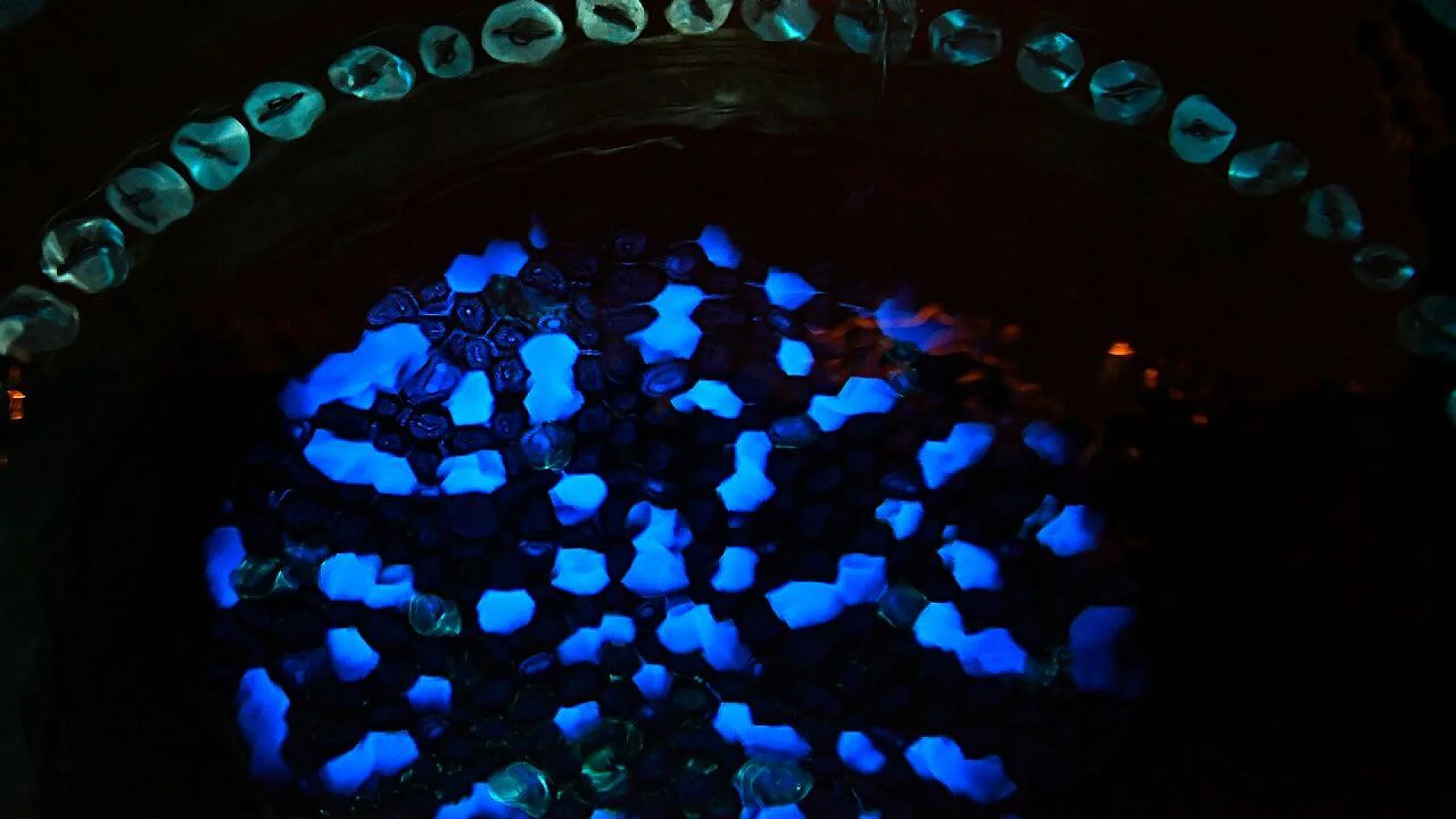 A cluster of luminous, crystalline blue shapes glowing intensely against a pitch-black background, framed by a curved line of translucent beads. The light resembles the ethereal blue glow of Cherenkov radiation.