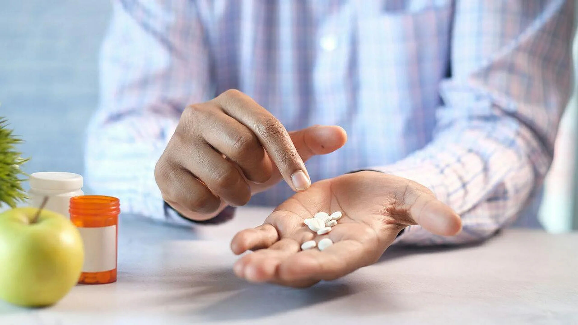 A close-up photograph shows a person holding several white, round supplement tablets in their open palm. An orange pill bottle with a white cap and a green apple sit on the table in the blurred background.