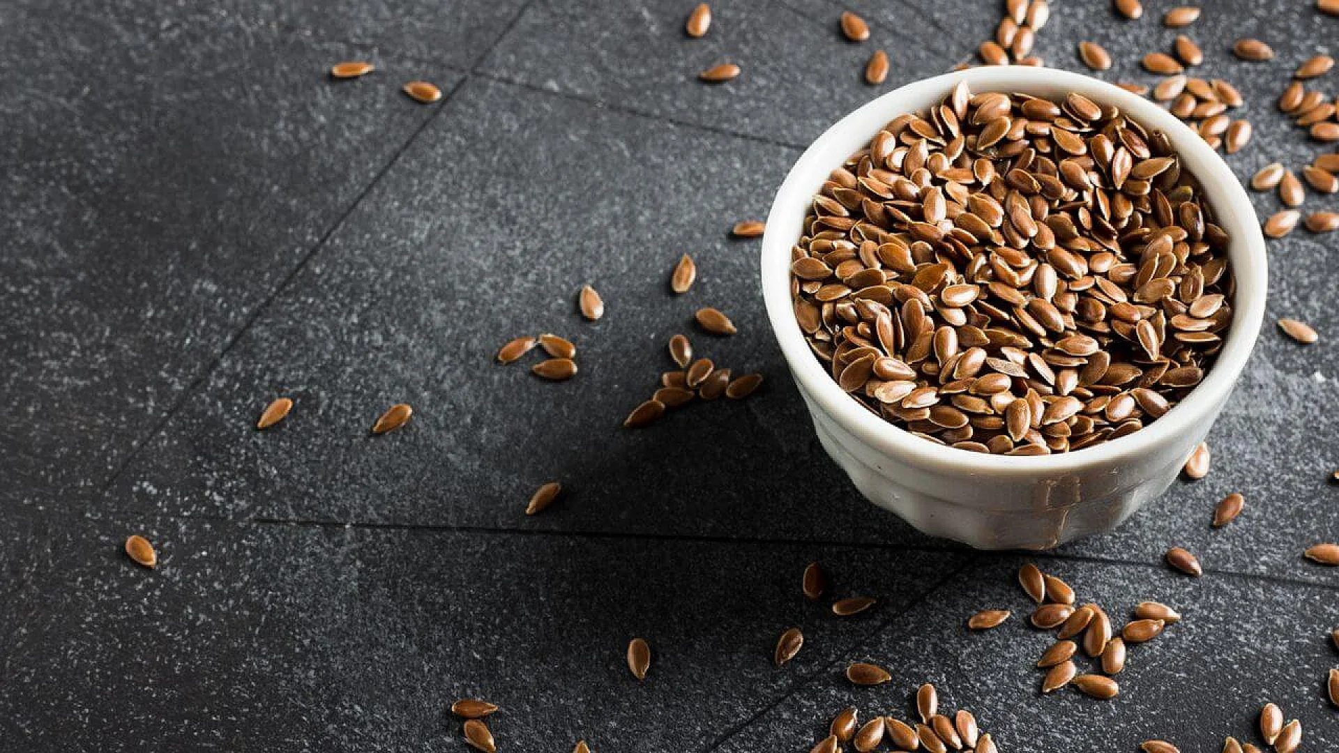 Close-up of whole brown flaxseeds in a small bowl, with some seeds scattered on a dark background.
