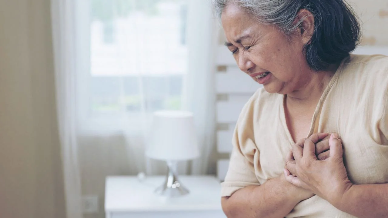 An older woman with grey hair winces in pain and clutches her chest with both hands, illustrating the sudden onset of symptoms in cardiovascular disease.