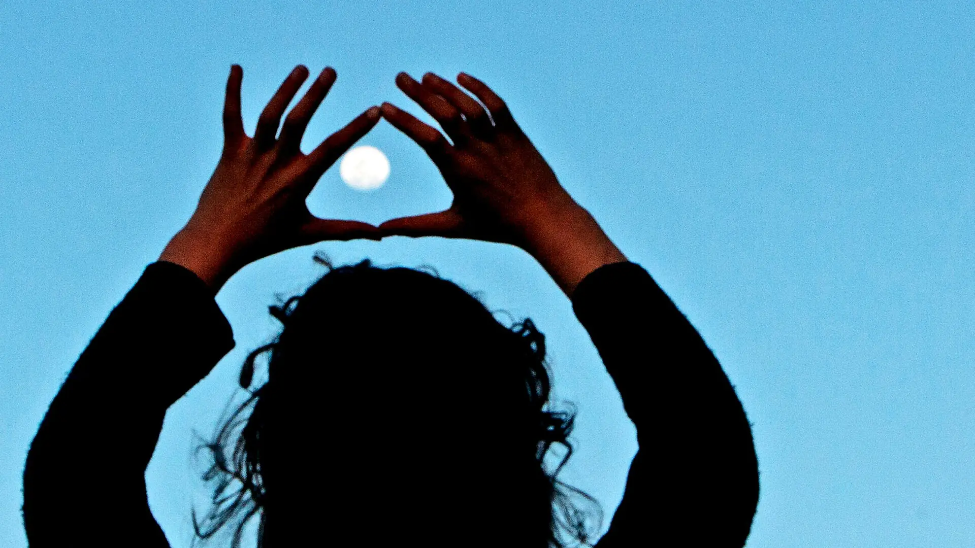 A woman holds her hands up to frame the moon in the evening sky.
