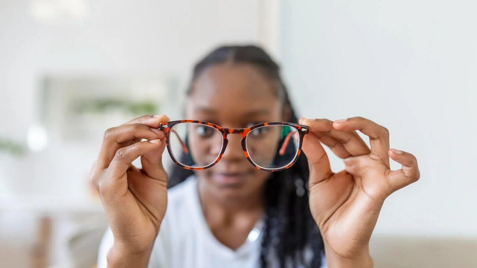 A person’s hands hold a pair of brown tortoise-shell eyeglasses in front of their face. The background is blurred, and the clear lenses of the glasses are the central focus.