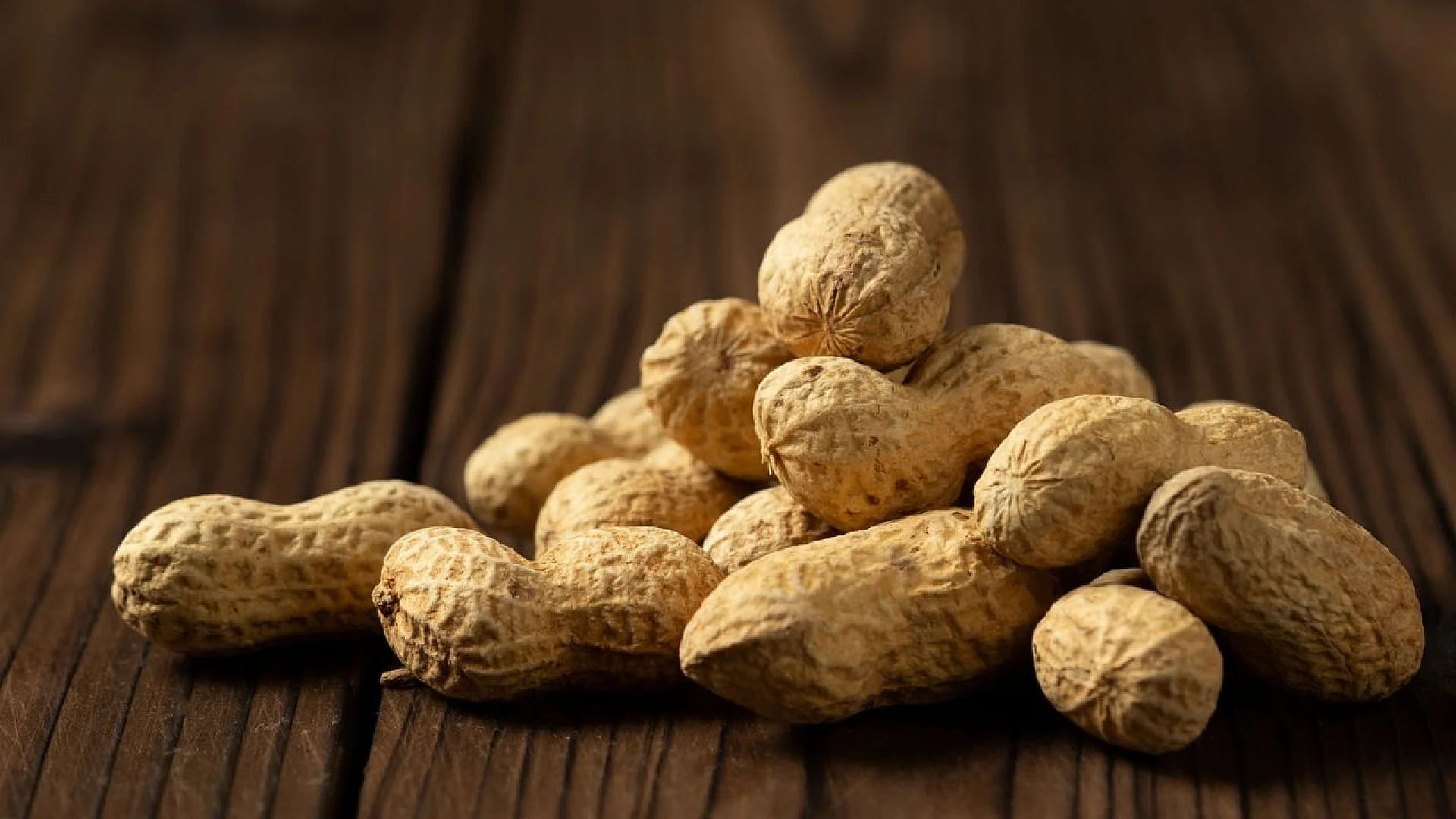 A close-up, shallow-depth-of-field shot of a pile of whole peanuts in their shells resting on a rustic, dark wooden surface.