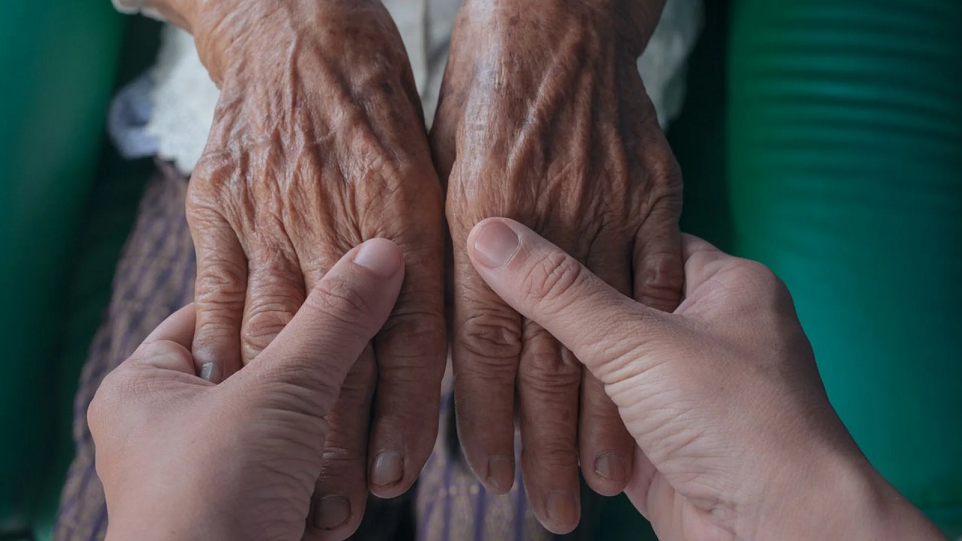 A close-up, top-down view of a younger person’s hands gently holding and comforting the wrinkled, sun-spotted hands of an elderly individual.