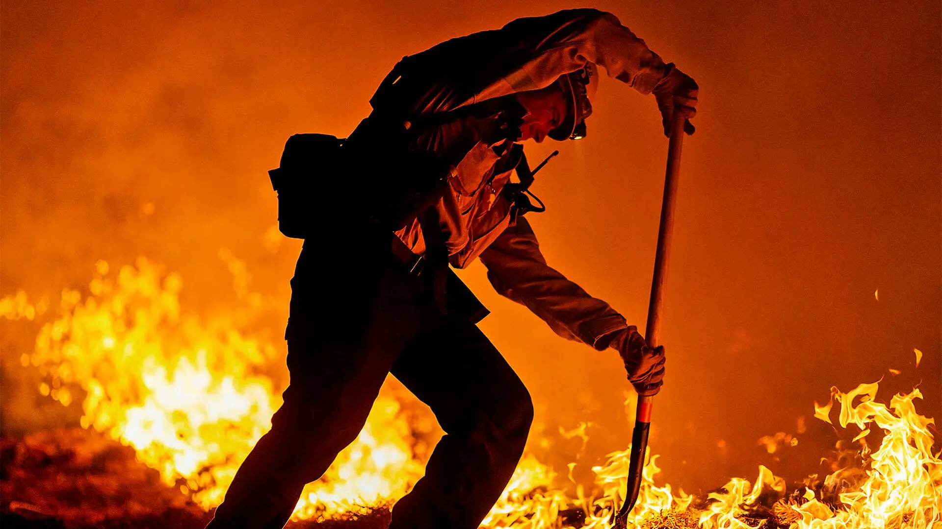 A firefighter uses a shovel to secure a fire break while flames burn behind him and the air is bright orange.
