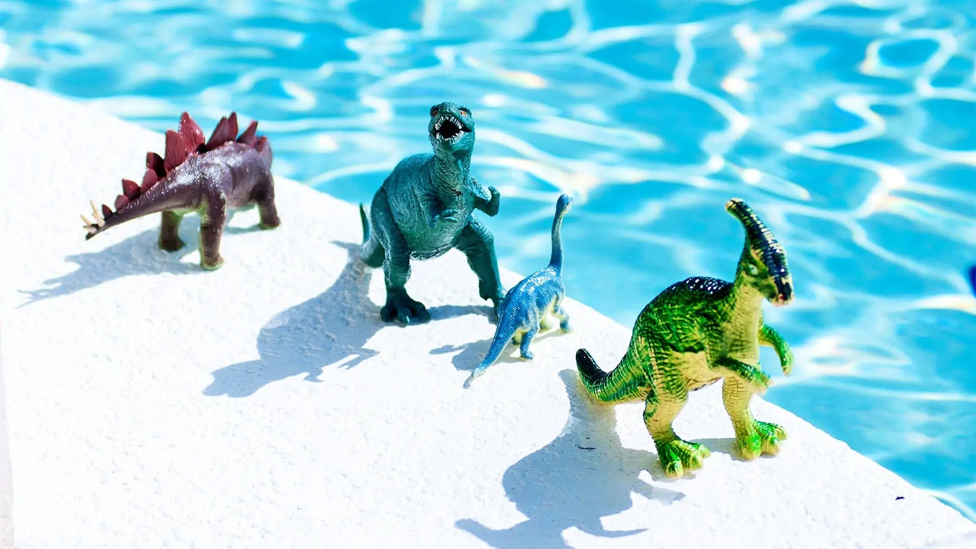 Dinosaur toys stand on the side of a blue swimming pool.