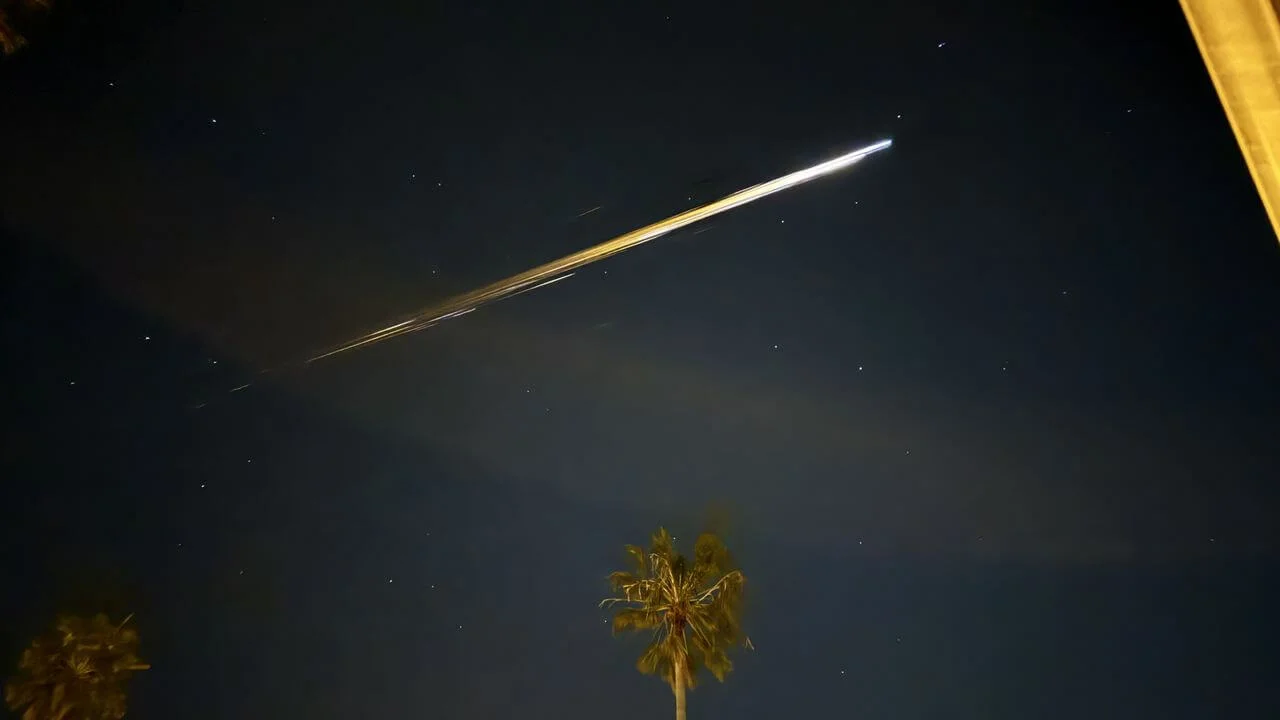 A night sky photograph showing a bright, diagonal orange light streak from a reentering spacecraft above a row of silhouetted palm trees in Ventura, California.