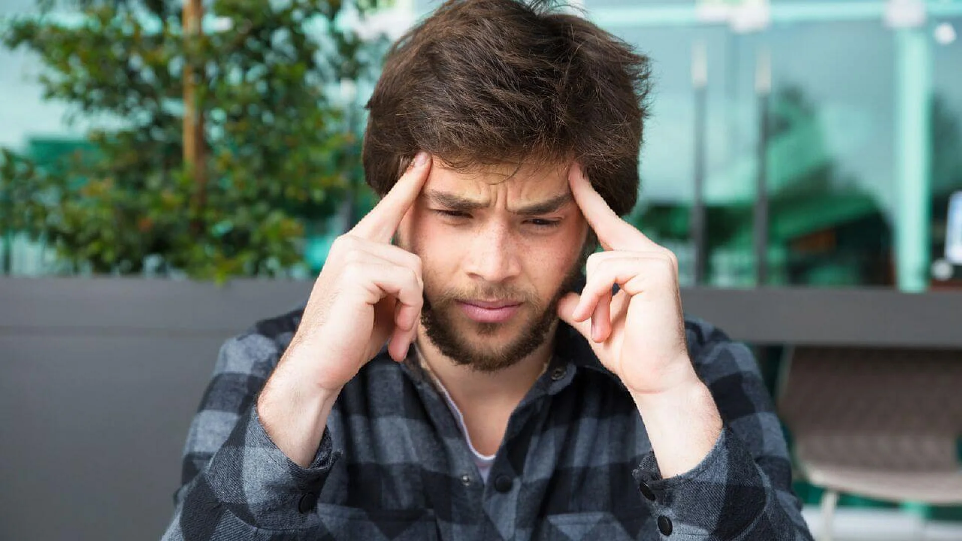 A young man with a beard looking down and pressing his fingers to his temples in deep concentration or frustration against a blurred modern building background.