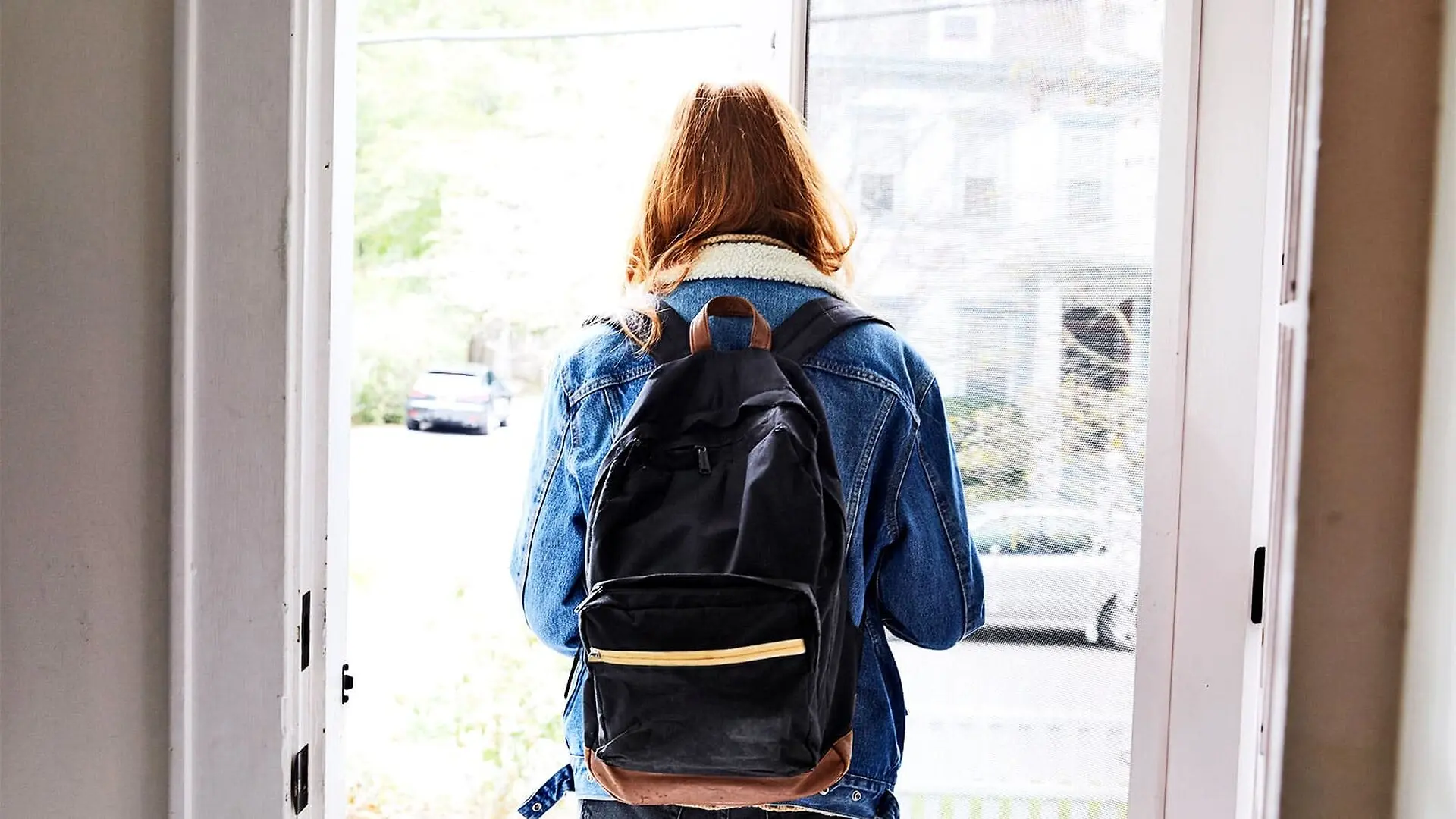 A teen girl walks out a front door alone while wearing a backpack.