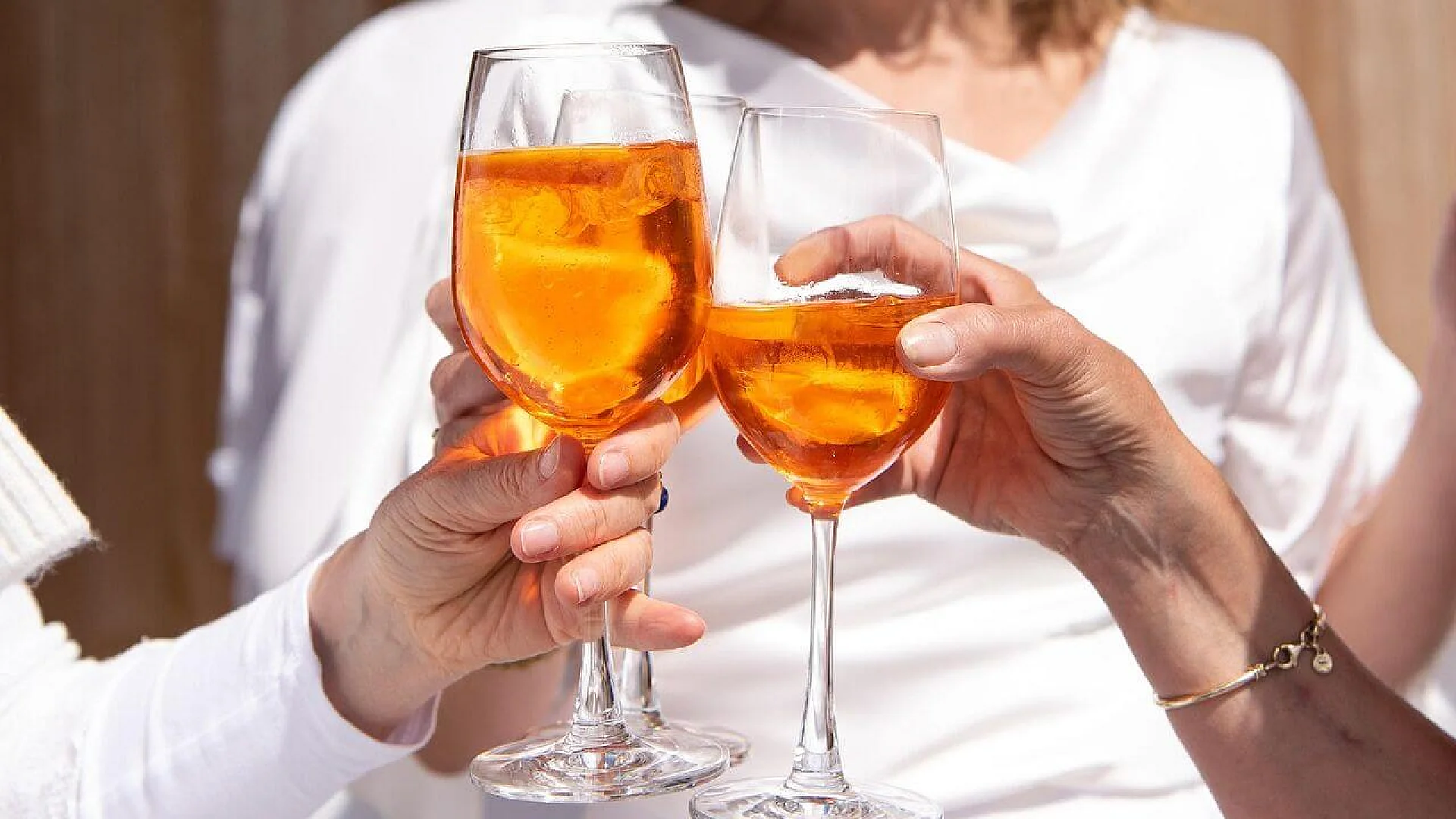 Two people in white shirts clinking glasses filled with orange cocktails (Aperol Spritz) during a daytime social gathering.