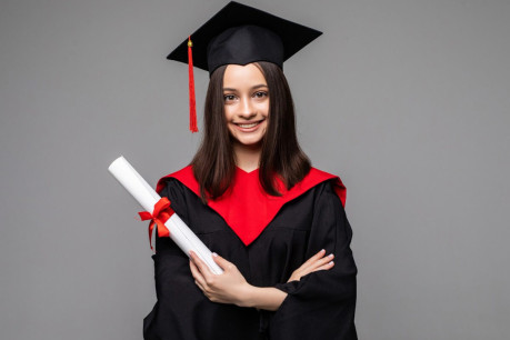 Happy student with graduation hat and diploma on grey background.